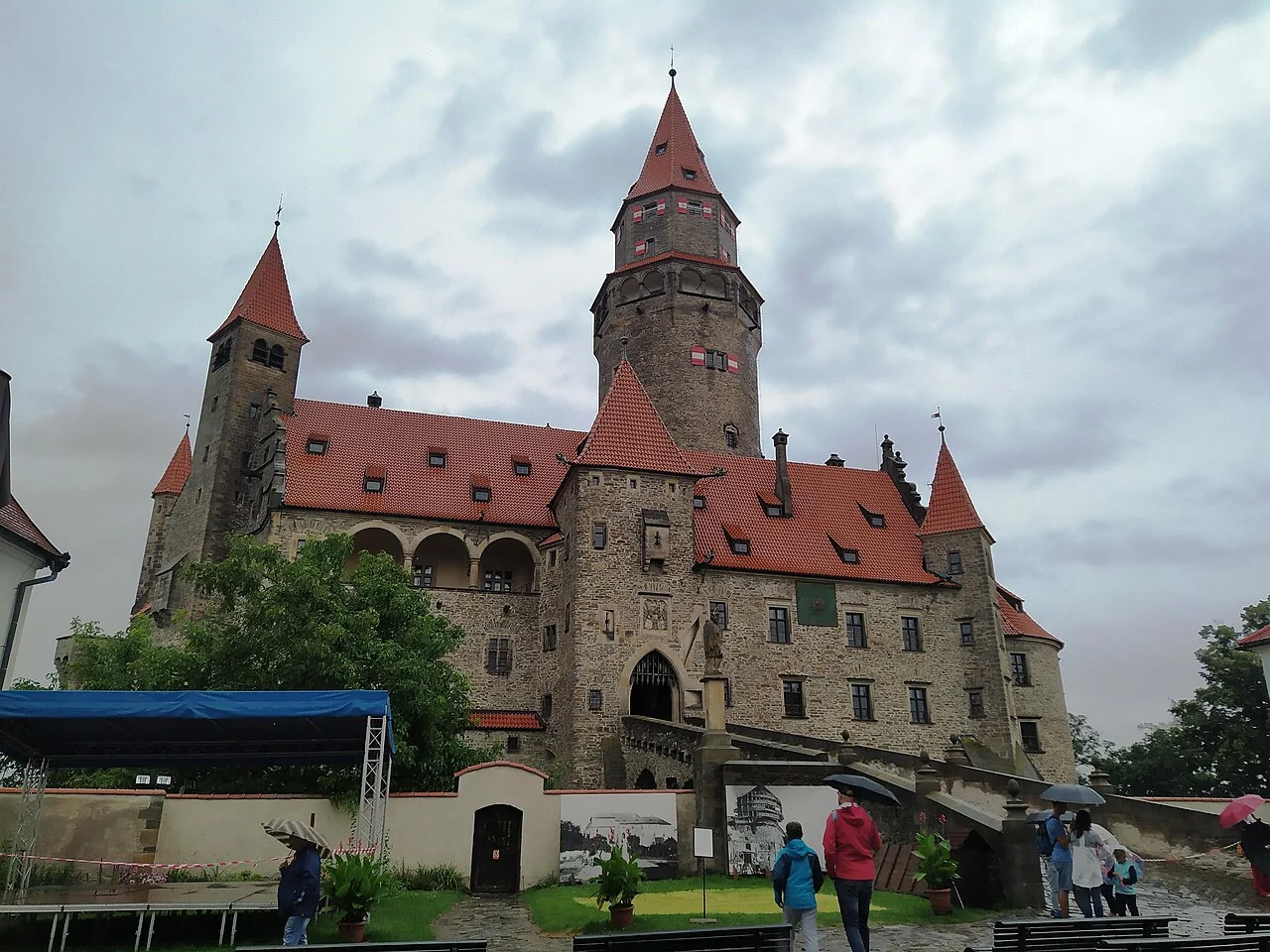 Hrad Bouzov – Celkový exteriér hradu Bouzov včetně typických věží; popis a souřadnice konkrétního souboru potvrzují Bouzov. V popředí návštěvníci a dočasné vybavení.